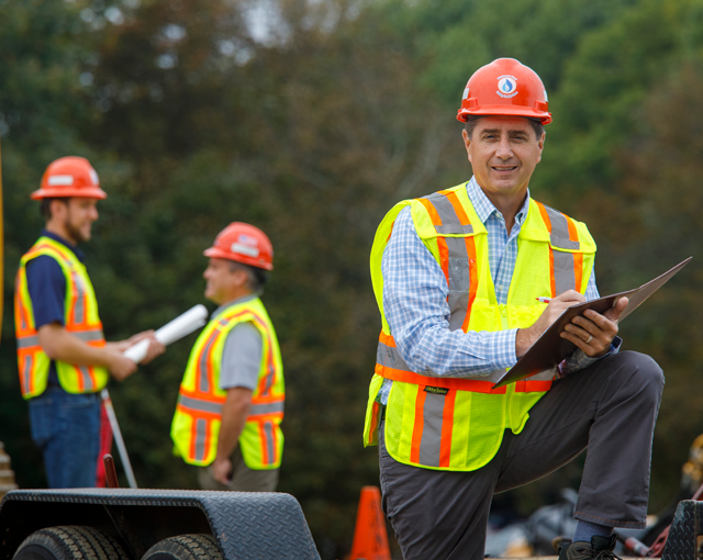 man with paper and pen in hard hat standing in front of two men talking in background