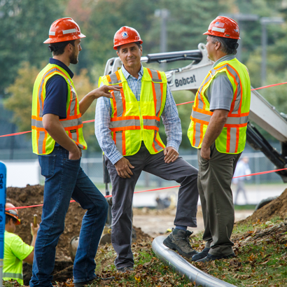 three men in hard hats and vests talking in front of machinery