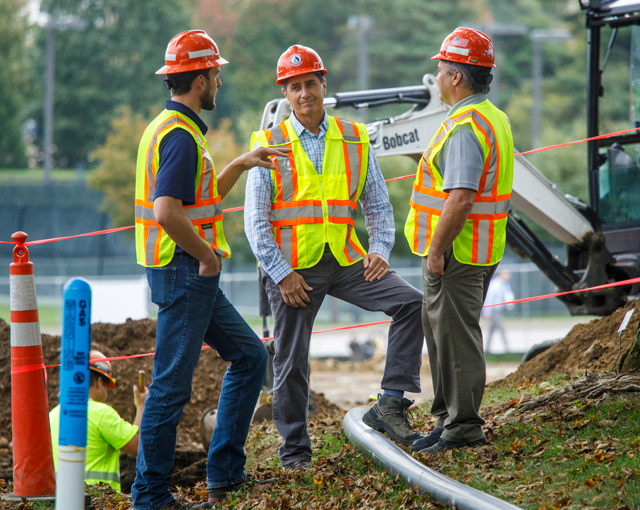 three men in hard hats and vests talking in front of machinery