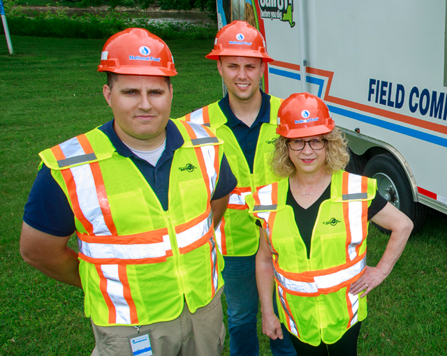 group of three people in hard hats and vests in front of field command center trailer