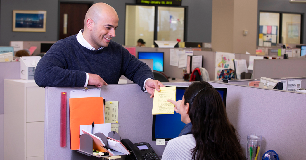 man holding a notepad talking to a woman in a cubicle