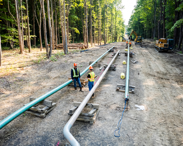 two men amid a worksite standing in between two pipes